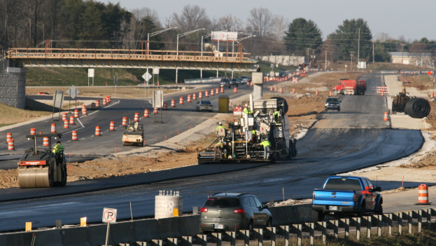 Baustelle mit Fahrzeugen auf der Straße, Verkehrskegel, Hinweisschilder, Pfosten, eine Brücke, Bäume und einen klaren blauen Himmel.