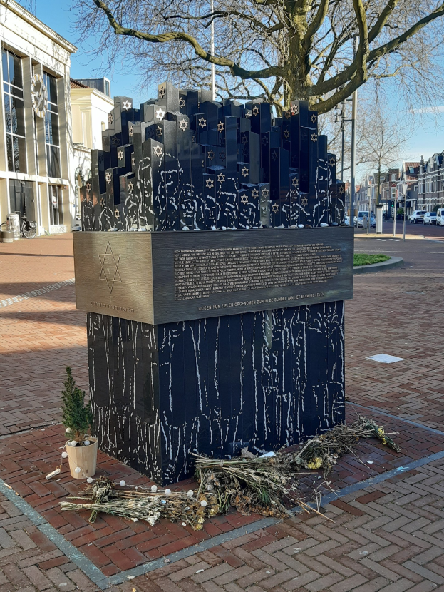 Mahnmal für Holocaust-Opfer in einem Stadtplatz mit einem Baum davor, Topfpflanze, verstreute trockene Blätter, umgeben von Gebäuden, Fahrzeugen, Pfählen, Bäumen und Himmel.