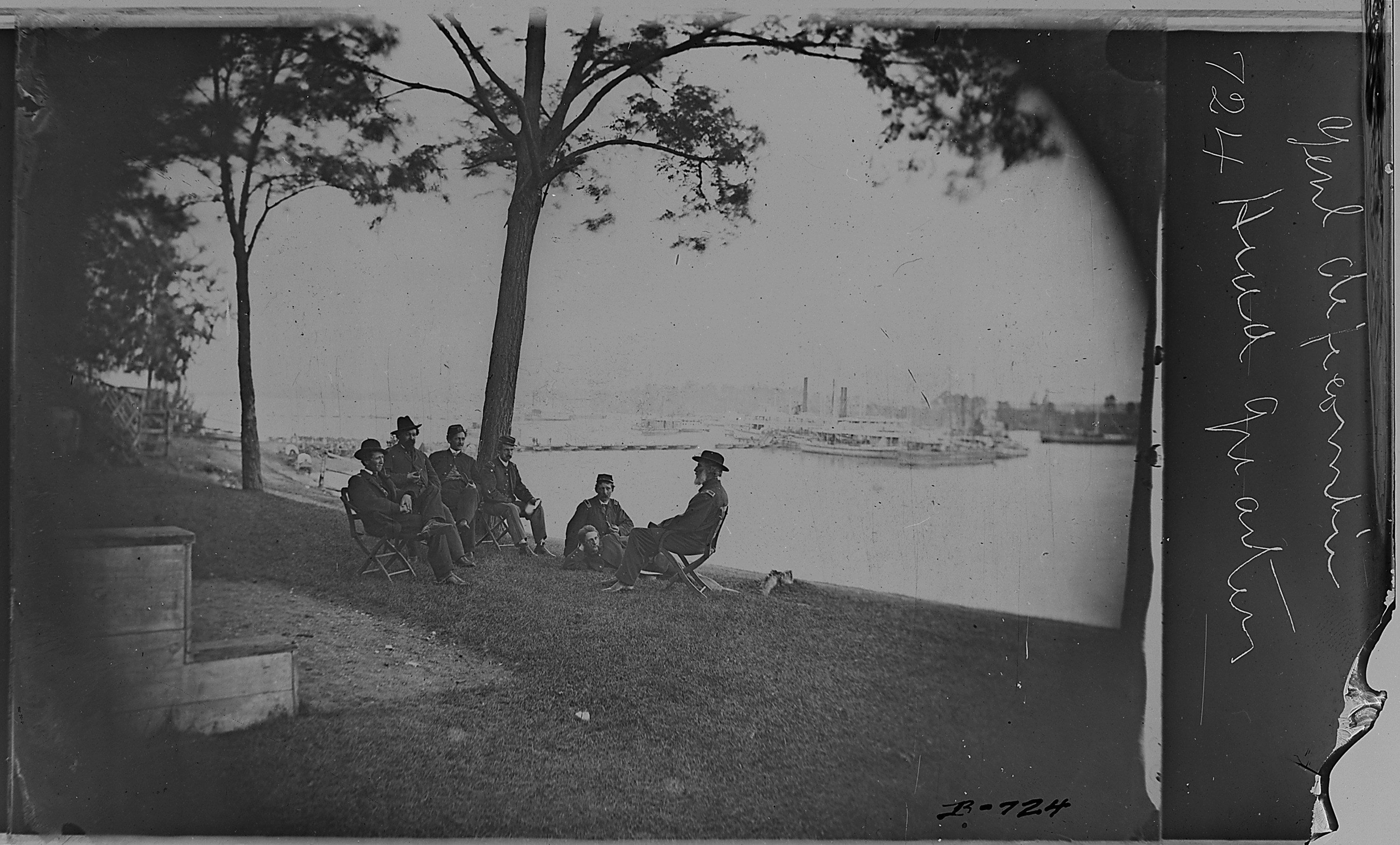 Eine Gruppe von Männern sitzt auf einer grünen Wiese und trägt Hüte, umgeben von Bäumen, mit Booten auf einem Gewässer im Hintergrund und Himmel darüber, mit Text auf der rechten Seite.