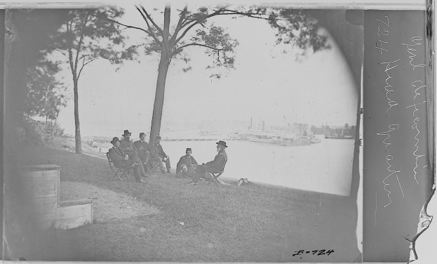 Eine Gruppe von Männern sitzt auf einer grünen Wiese und trägt Hüte, umgeben von Bäumen, mit Booten auf einem Gewässer im Hintergrund und Himmel darüber, mit Text auf der rechten Seite.