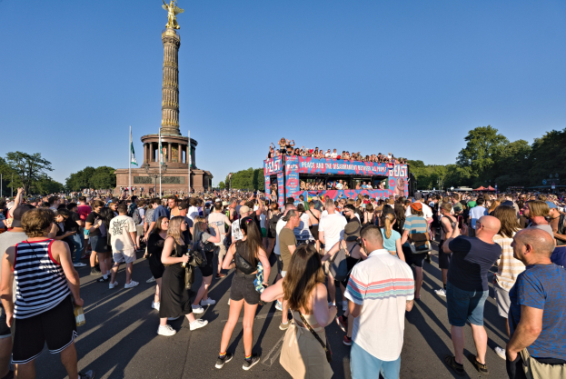 Große Menschenmenge versammelt sich vor einem Denkmal in Berlin mit Fahnen, einem säulengeschmückten Gebäude mit Statue und Inschrift, Bäumen und einem klaren blauen Himmel.