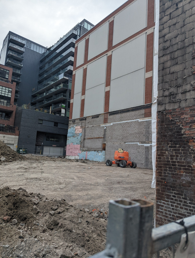 Baustelle mit einem großen Gebäude unter Abriss, einem Fahrzeug, einem Metallzaun und einem bewölkten Himmel.