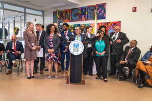Gruppe von Menschen vor einem Rednerpult mit Mikrofon und einer Tafel mit Text, mit sitzenden und stehenden Teilnehmern, Flaggen, Feuerlöscher und Deckenleuchten im Hintergrund.