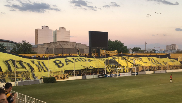 Ein Fußballfeld mit einer Gruppe von Menschen darauf, umgeben von einem Metallzaun, einem Tor, einer Fahne mit der Aufschrift "Boca Juniors vs. Real Sociedad", einigen Pflanzen, einem Anzeigebildschirm, einigen Pfosten, der Rinde eines Baums, einigen Bäumen, einigen Gebäuden und einem bewölkten Himmel.