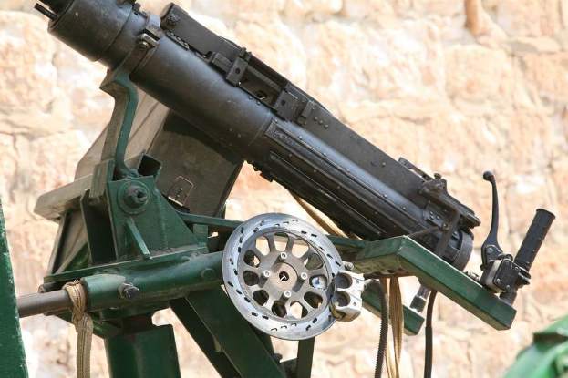Eine Maschinengewehr auf einem Stahlständer mit einem Griff auf der rechten Seite, Seilen und Kabeln unten und einer Wand im Hintergrund.