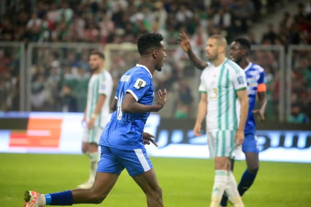 Ein Fußballspieler in einem blauen und weißen Trikot feiert mit erhobenen Armen, umgeben von einer Gruppe von Menschen in weißen und grünen Trikots, mit einem unscharfen Hintergrund, der ein weißes und blaues Brett und ein Geländer zeigt.