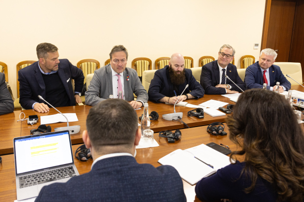 Eine Gruppe von Menschen, wahrscheinlich Mitglieder des Europäischen Parlaments, sitzt um einen Tisch in einem Konferenzraum, umgeben von Laptops, Bücher, Papieren, Flaschen, Gläsern, Headsets, Mikros mit Ständern und Mobiltelefonen, mit einer Wand und einer Tür im Hintergrund.