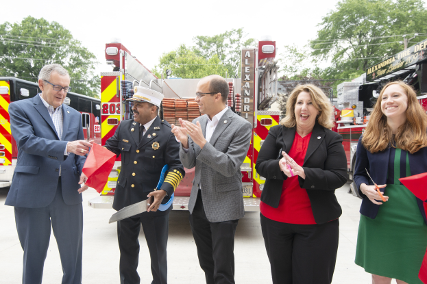 Eine Gruppe von Menschen steht vor einem Löschfahrzeug, klatscht und lacht, mit zwei Personen, die eine Schere und ein rotes Tuch halten, während einer Einweihungszeremonie für die Alexandria Fire Department, mit Bäumen, Drähten und einem klaren blauen Himmel im Hintergrund.