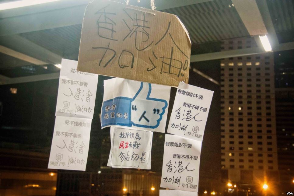 Menschen vor einem beleuchteten Gebäude bei Nacht mit Protestschildern an der Decke.