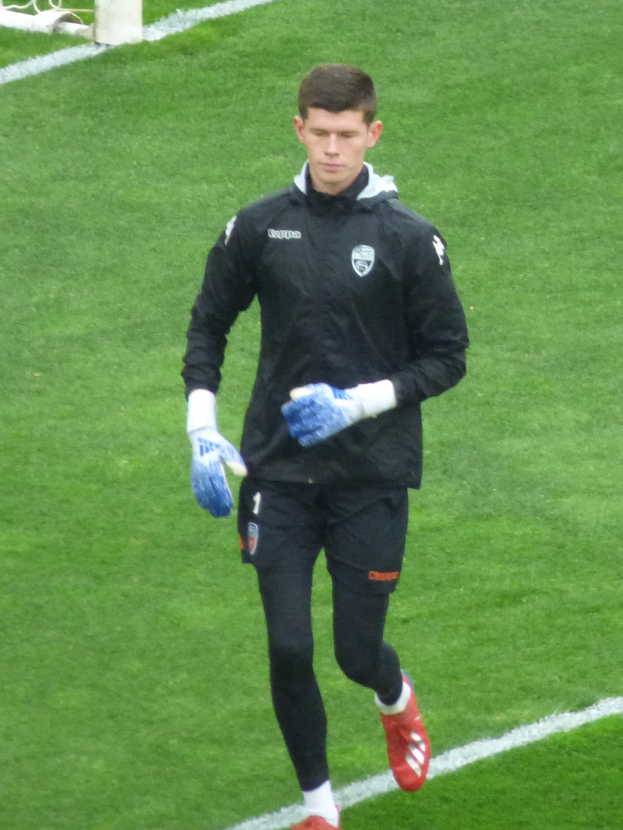 Ein Mann in einer schwarzen Jacke und blauen Handschuhen geht auf einem Fussballfeld mit einem Tor im Hintergrund, wobei er Fussballhandschuhe tr├Ągt.