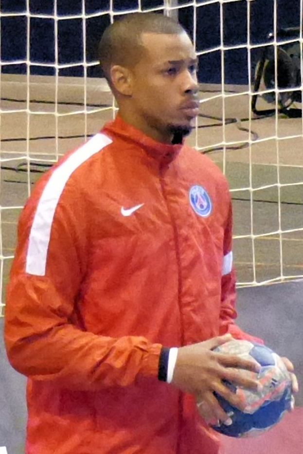 Ein Mann in einem blauen Hemd und Shorts steht vor einem Fußballtor und hält entschlossen einen Ball.