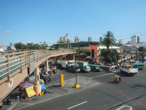 Vielbefahrene Straße mit starkem Verkehr, eine Brücke, Fußgänger, Zelte, Versorgungsmasten, Verkehrsampeln, Bäume, Gebäude, Wasser im Hintergrund und ein bewölkter Himmel.