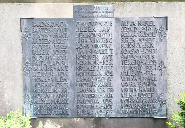 Eine Steinplatte an einer Wand mit eingraviertem Text, umgeben von Pflanzen am Boden, wahrscheinlich ein Holocaust-Mahnmal.