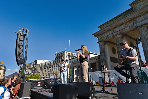 Eine Gruppe von Menschen auf der Bühne vor dem Brandenburger Tor in Berlin, Deutschland, die Musikinstrumente spielen und in Mikrofone singen, mit Lautsprechern und anderen Gegenständen drumherum, sowie Gebäuden, Bäumen, Pfählen, Flaggen und einem klaren blauen Himmel im Hintergrund.