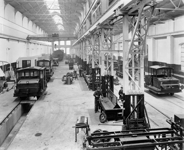 Schwarzes und weißes Foto einer Fabrikhalle mit mehreren reparierten Zügen, Arbeitern, die sich darum kümmern, Metallstützen, Deckenlampen und Wandfenstern.