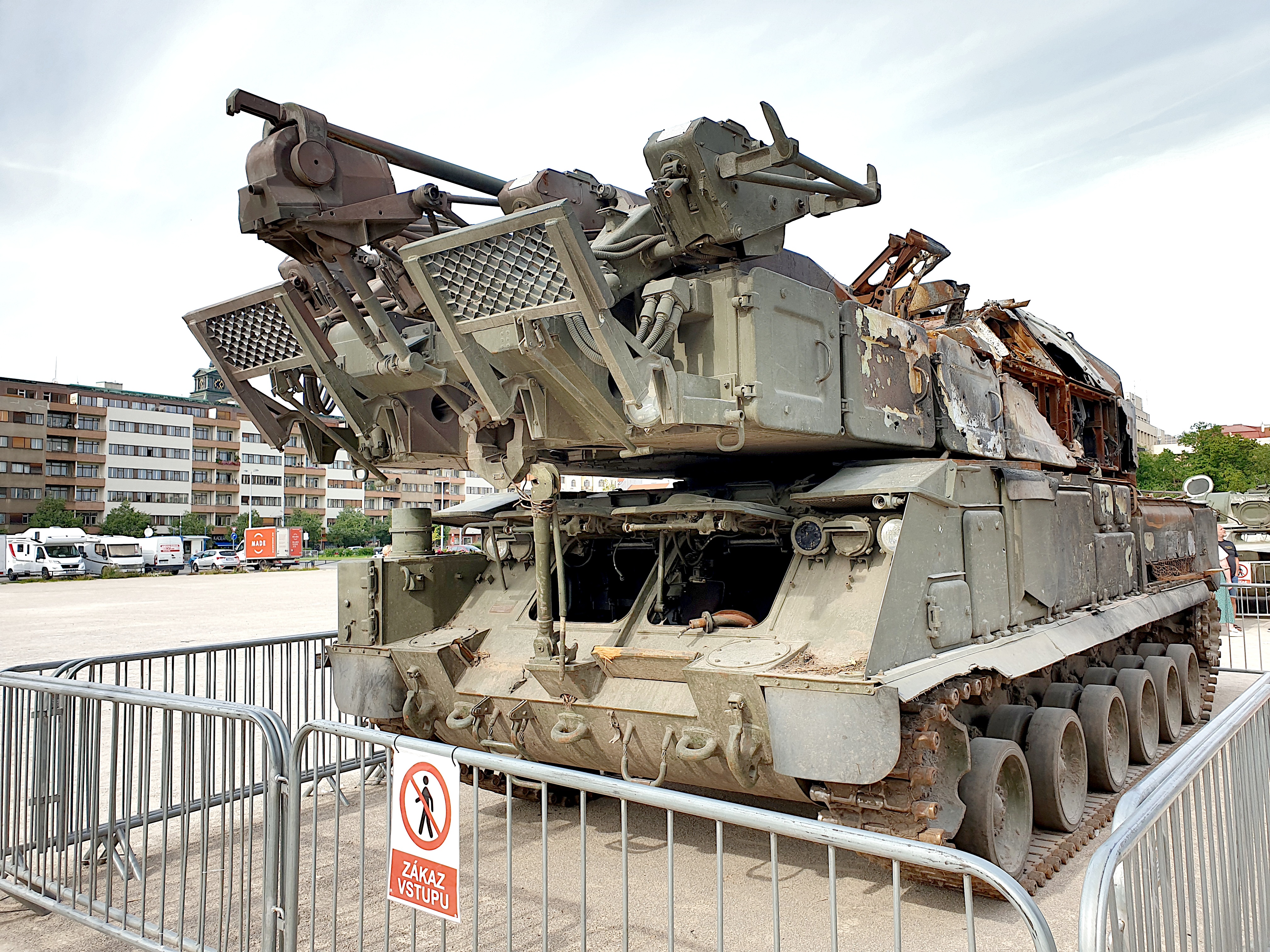 Ein gepanzerter Mannschaftswagen steht vor einem Metallzaun mit einem daran befestigten Schild, mit Fahrzeugen, Bäumen, Gebäuden und einem klaren blauen Himmel im Hintergrund.