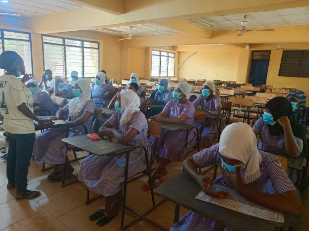 Ein Klassenzimmer in Nigeria mit Schülern, die Gesichtsmasken tragen und auf Bänken und dem Boden sitzen, mit Büchern und anderen Gegenständen auf den Tischen, Fenstern, einer Tür, einer Wand, einer Decke mit Lampen und Ventilatoren im Hintergrund.