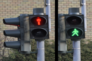 Zwei Ampeln an einem Mast mit roter und grüner Leuchte, vor einer Backsteinwand und Bäumen positioniert, die einen Fußgängerübergang anzeigen.