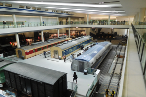 Ein großer Museumsraum mit verschiedenen Bahnen in unterschiedlichen Größen und Farben, mit Menschen, Geländern, Säulen und Deckenleuchten.
