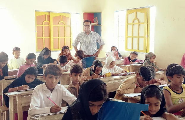 Eine Gruppe von Kindern sitzt an Tischen in einem Klassenzimmer in Pakistan, mit Büchern und Stiften in den Händen, mit einer Person im Hintergrund und einem büchergefüllten Schrank im Hintergrund und Fenstern an den Wänden.