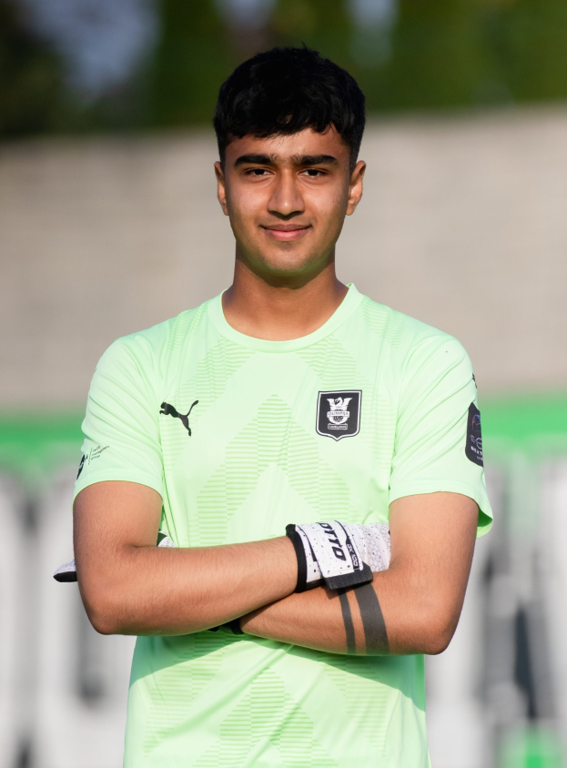 Ein junger Mann in einem grünen T-Shirt steht mit verschränkten Armen auf einem Fußballfeld und lächelt, mit einem unscharfen Hintergrund.