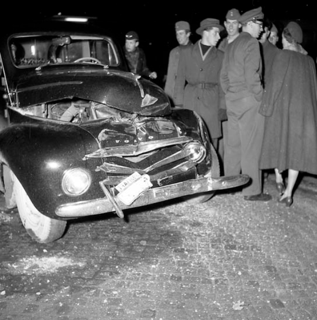 Ein Schwarz-Weiß-Bild eines auf der Straße liegenden, stark beschädigten Autos mit einer Gruppe besorgter Menschen drumherum.