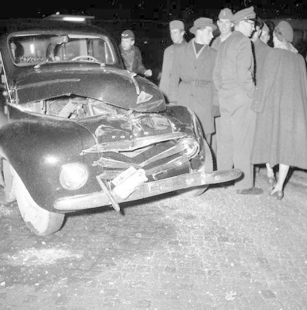 Schwarz-weißes Bild eines schwer beschädigten Autos am Straßenrand, mit einer Gruppe von Menschen drumherum, die die eingedrückte Motorhaube und die zersplitterten Fenster untersuchen.