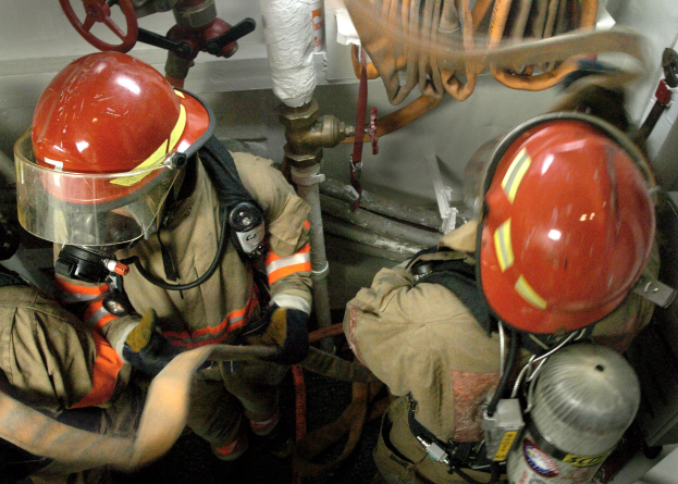 Feuerwehrleute in Helmen und Schutzausrüstung arbeiten an einem Feuerhydranten mit Rohren und Ventilen im Hintergrund.