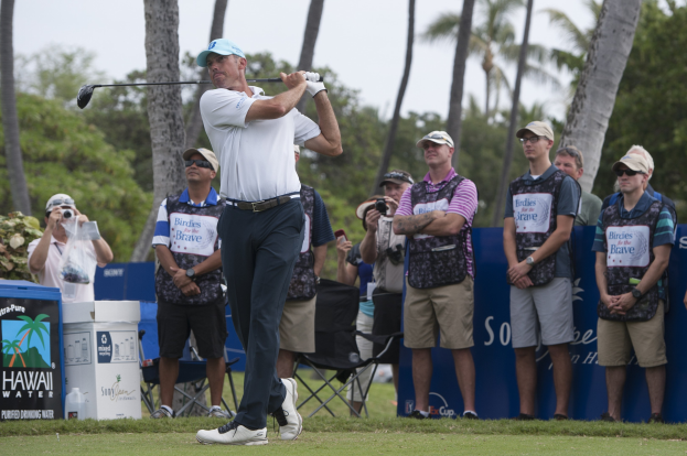 Ein Mann in einem weißen T-Shirt, schwarzen Hosen und einer blauen Mütze schwingt einen Golfschläger auf einem saftig grünen Feld, mit Zuschauern und Fotografen dahinter, vor einem Hintergrund aus Bäumen und einem klaren blauen Himmel.