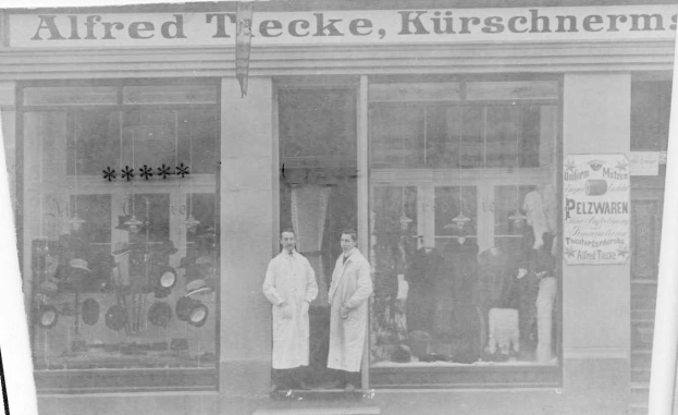 Zwei Männer stehen vor einem Geschäft in Berlin, Deutschland, mit einem Schild, auf dem "Alfred Tecke, Kurschnerm" steht, während Schaufensterpuppen in Kleidern und Hüten zu sehen sind und ein Textbrett auf der rechten Seite.