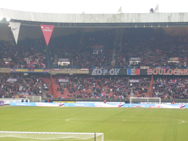 Ein grasbewachsener Stadionboden mit Torpfosten auf beiden Seiten, Zuschauern im Hintergrund und zahlreichen Werbetafeln in der Mitte und darüber.