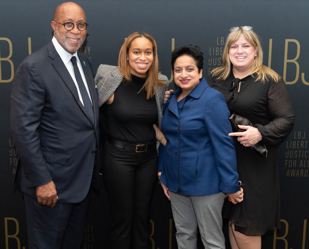 Eine Gruppe von vier Menschen, die auf einem roten Teppich stehen, lächeln und für ein Foto posieren, mit einem Schild im Hintergrund, auf dem "Freiheit Gerechtigkeit für alle Auszeichnungen" steht.