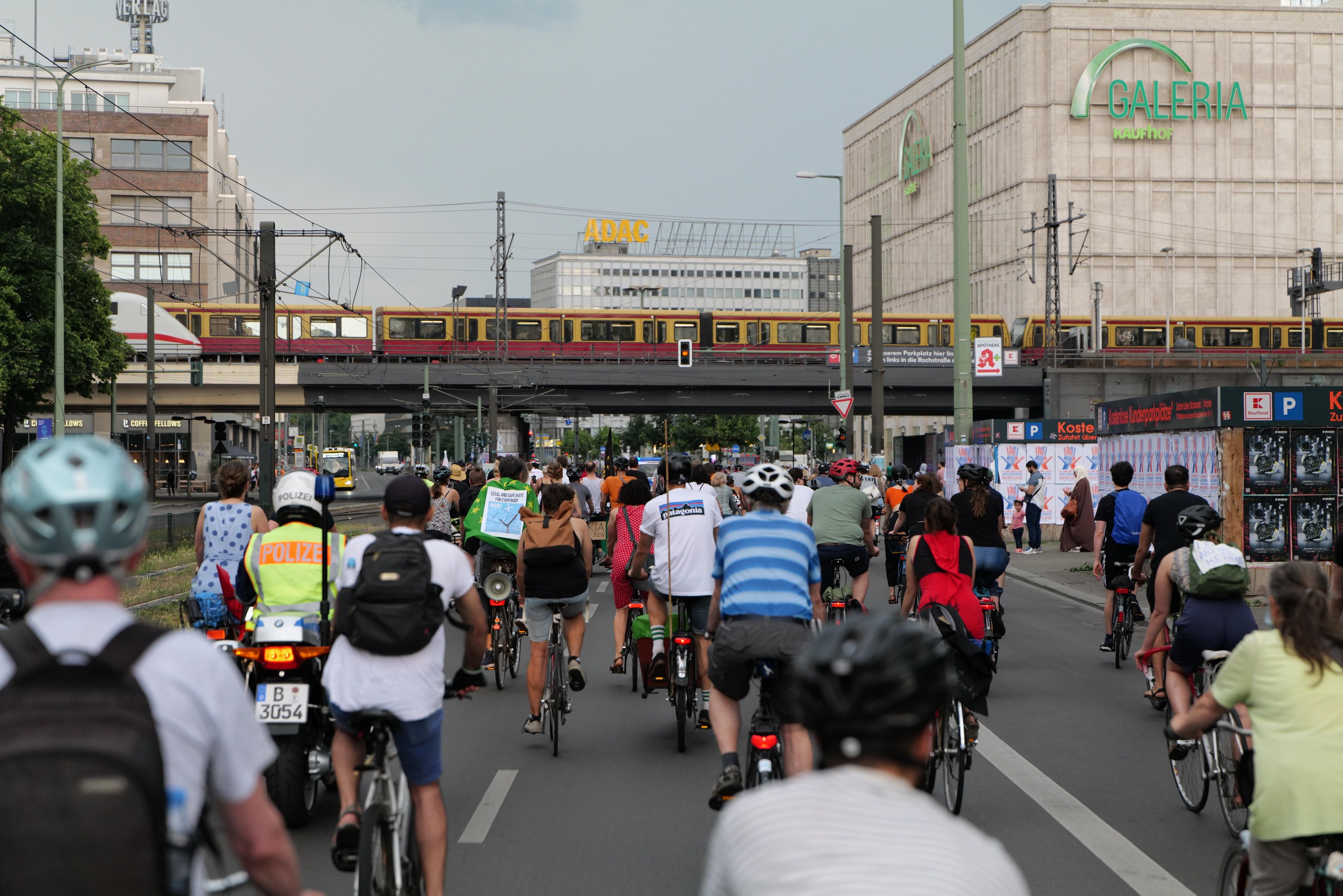 Eine Gruppe von Menschen, die auf Fahrrädern eine Straße entlangfahren, die von hohen Gebäuden gesäumt ist, einige tragen Helme und Taschen, mit einem Zug auf einem Bahngeleis, Strommasten, Bäumen und einem klaren blauen Himmel im Hintergrund.