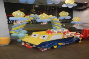 Eine McDonald's-Geburtstagsparty-Szene mit einem gelben und blauen Lastwagen, der mit Ballons geschmückt ist, einem rot gedeckten Tisch mit Gegenständen, Hockern und einer Glaswand im Hintergrund unter festlicher Deckenbeleuchtung.