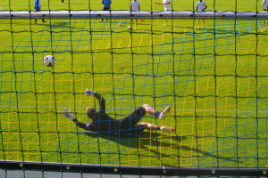 Ein Fußballspieler taucht ab, um einen Ball vor einem Tor zu halten, mit einem Netz im Vordergrund und ein paar Menschen im Hintergrund, während mehrere Fußballs auf dem Boden verstreut sind.