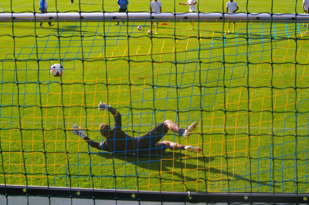 Ein Fußballspieler taucht ab, um einen Ball vor einem Tor zu halten, mit einem Netz im Vordergrund und ein paar Menschen im Hintergrund, während mehrere Fußballs auf dem Boden verstreut sind.