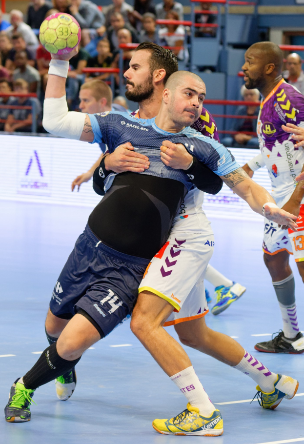 Gruppe von Männern beim Handballspielen in einem Stadion, einer hält den Ball, mit Zuschauern im Hintergrund.