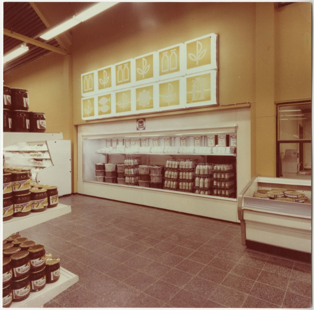 Alte Fotografie eines Ladeneinrichtungsinneren mit Regalen voller Lebensmittelbehälter, einem Kühlschrank, einer Wandtafel, Deckenleuchten und einem Fenster.