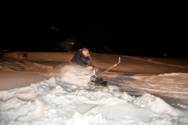 Eine Person beim Skifahren auf dem Schnee mit einer weiteren Person im dunklen Hintergrund.