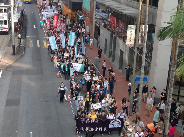 Eine große Gruppe von Menschen marschiert mit Protestschildern eine Straße entlang, mit Bäumen und Glasgebäuden auf der rechten Seite, Fahrzeugen auf der linken Seite und Straßenschildern und Pfählen.