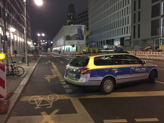 Ein Polizeiwagen steht nachts an der Seite einer Straße in Berlin, Deutschland, umgeben von Gebäuden, Straßenlaternen, Motorrädern, Absperrpollern, Fahrrädern, Menschen auf dem Gehweg, Werbetafeln und einer Statue im Hintergrund, wahrscheinlich nach einer Schießerei und einem Autounfall.