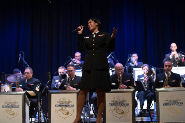 Frau in militärischer Uniform, die ein Mikrofon auf einer Bühne hält, umgeben von sitzenden Musikern, die Instrumente spielen, mit Mikrofonen und einem Vorhang im Hintergrund.