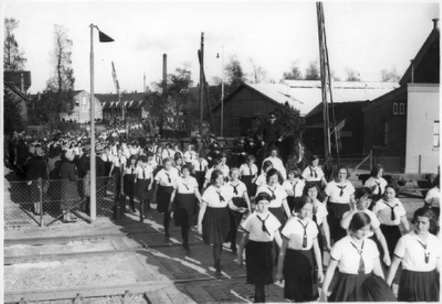 Eine Gruppe von Mädchen in Schuluniformen marschiert die Straße entlang, mit einem Zaun auf der linken Seite und Menschen dahinter, Pfählen, Bäumen und Gebäuden im Hintergrund und einem sichtbaren Himmel, in Schwarz-Weiß.