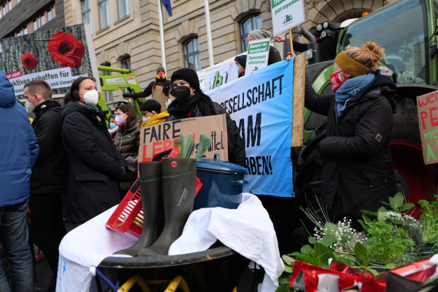 Eine Gruppe von maskierten Menschen mit Protestschildern vor einem Lastwagen, mit einem Tisch, Pflanzen, Gebäuden und einer Fahne im Hintergrund.