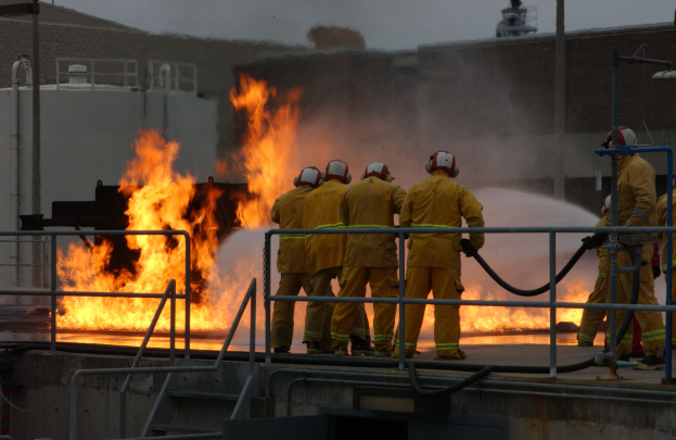 Feuerwehrleute in Helmen stehen auf einem Gebäudedach und halten Rohre, mit Geländern und Treppen darunter und einem Gebäude und Himmel im Hintergrund.