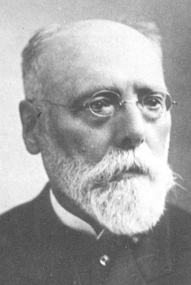 Schwarzes und weißes Foto eines Mannes mit weißem Bart, der ein schwarzes Kleid und eine Brille trägt und ernst schaut.