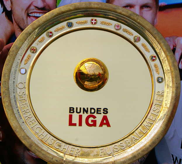 Mann hält goldenes Schild mit "Bundesliga" hoch, im Hintergrund ein Banner mit drei Personen