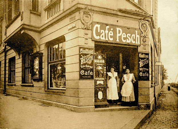 Ein altes Schwarz-Weiß-Foto von zwei Frauen vor einem Café mit Fenstern, einer Tür und Schildern, mit Bäumen und einem klaren Himmel im Hintergrund.