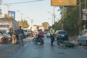 Gruppe von Menschen um ein verunglücktes Motorrad am Straßenrand mit mehreren Fahrzeugen, darunter ein Lastwagen, im Hintergrund und Bäumen, Pfählen, Lampen, Schildern und Himmel.