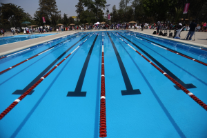 Großer Schwimmbad mit Bahnteiler, umgeben von Menschen mit Taschen und Gegenständen, sowie Zelten, Schildern, Stangen und einem bewölkten Himmel im Hintergrund.